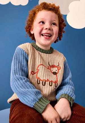 Vörös hajú gyerek bézs és kék kötött pulóvert visel, amelyen egy rajzfilm tehén látható, mosolyogva egy kék fal előtt, amelyen fehér felhő dekorációk vannak.