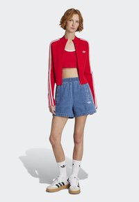 Rote Zip-Jacke mit weißen Streifen, rotes Crop-Top, Jeansshorts und weiße Sneakers mit schwarzen Akzenten. Weiße Socken mit Logodetail.