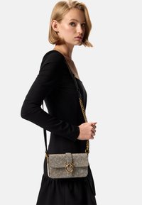 Piccola borsa strutturata in tessuto argentato texturizzato con un accento in metallo dorato e una tracolla a catena, indossata a tracolla. Sfondo di un abito nero.