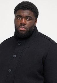 Black wool blend coat featuring a high collar, button front, and side pockets. Structured design with subtle stitching details.