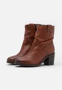 Para brązowych skórzanych botków do połowy łydki z bocznym zamkiem błyskawicznym, zastawioną blokową podeszwą, okrągłymi czubkami i dekoracyjnym przeszyciem na białym tle.