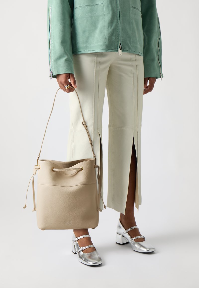 Bežová kožená kabelka v tvare vedra so sťahovacím uzatváraním, hladkou textúrou a jemným brandingom. V kombinácii s mätovo zelenou semišovou bundou a svetlými nohavicami.