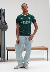 Groen voetbalshirt met patroon en "ETIHAD AIRWAYS" in het wit, gecombineerd met lichtblauwe spijkerbroek en grijze sneakers. Simpele kleedkamerachtergrond.