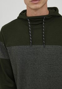 Kapuchon sweatshirt van donkergroene stof met een grijze paneel. Voorzien van zwart-witte trekkoord hardware. Zachte textuur en een relaxte pasvorm.