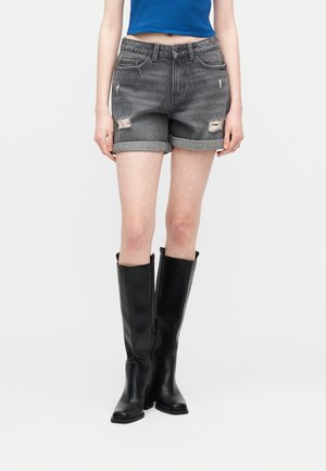 Kobieta ubrana w czarne, postarzałe szorty jeansowe, niebieski krótki top i czarne kozaki za kolano, stojąca na tle jednolitego białego tła.