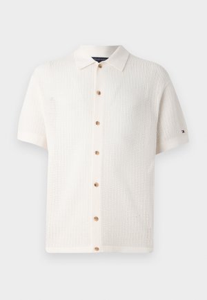 Polo shirt blanc à manches courtes en tricot, avec un col, présente une fermeture à boutons et un motif texturé. Logo de la marque sur la manche gauche.