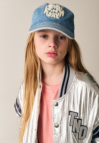 Denimkasket med broderet tekst, sølvfarvet bomberjakke med sort og hvidt detaljer, og lyserød t-shirt. Langt, lige hår.