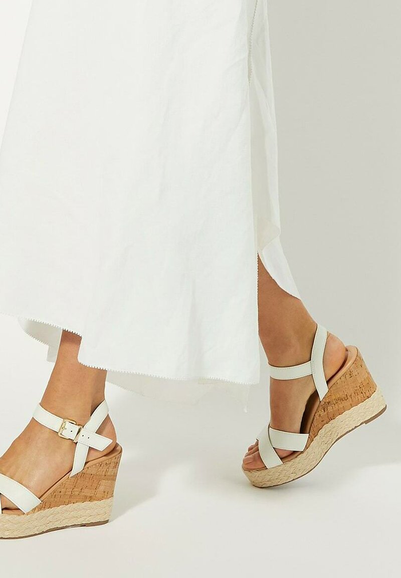 White wedge sandals featuring a cork platform, adjustable ankle strap, and smooth leather upper. The design includes two front straps for support.
