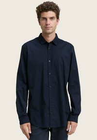 Uomo con capelli castani ricci che indossa una camicia a maniche lunghe blu scuro con bottoni, in piedi di fronte a uno sfondo chiaro e uniforme.