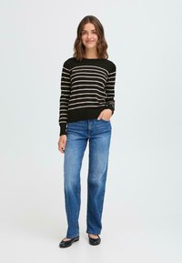 Maglione a strisce nere e beige, maniche lunghe, collo rotondo; abbinato a jeans blu a gamba dritta e scarpe nere basse. Sfondo semplice.