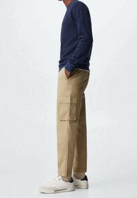 Maglione lavorato a maglia blu navy, pantaloni cargo beige con tasche laterali e sneakers bianche con dettagli neri. L'outfit presenta un design rilassato.