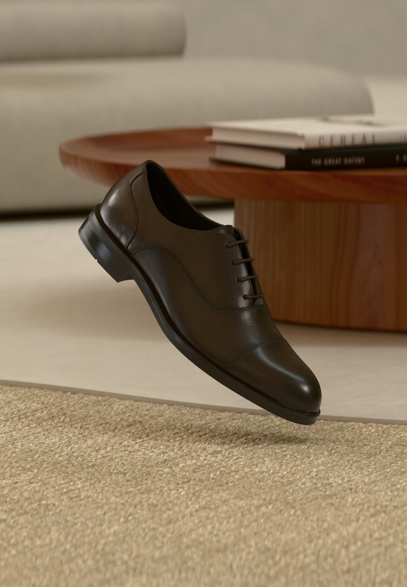 Chaussure Oxford en cuir noir flottant au-dessus d'un tapis tissé clair avec une table basse ronde en bois et des livres empilés en arrière-plan.
