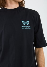 Sort bomulds t-shirt med en lys blå sommerfugl grafik og teksten "Overflod er en tankegang" nær brystet. Korte ærmer.