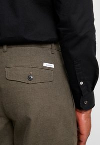 Olivově zelené kalhoty se zadní kapsou, na které je černý knoflík a bílá nálepka "Calvin Klein". Viditelná je černá dlouhá košile s dlouhým rukávem.