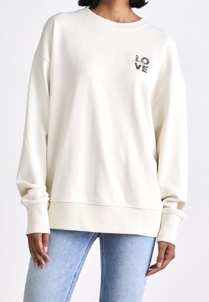 Persoon draagt een oversized crème sweatshirt met de tekst "LOVE" op de borst, gecombineerd met lichtblauwe spijkerbroek.