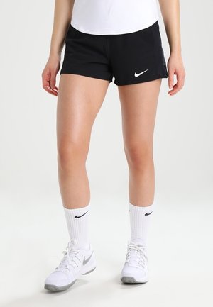 Kvinde iført sorte Nike-shorts, hvide Nike-strømper og hvide Nike-sneakers, der står mod en ensfarvet baggrund.