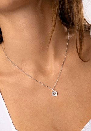 Silberne Halskette mit feiner Kette, die einen runden Anhänger mit dem Buchstaben "M" zeigt. Das Design ist minimalistisch und zeitgenössisch.