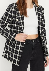 Mustavalkoinen ruudullinen bleiseri, jossa on teksturoitu kangas, kultaiset nappikoristeet ja muotoiltu leikkaus, yhdistettynä valkoiseen lyhytpaita.