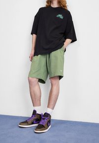 Svart oversized t-shirt med grön logotyp, ljusgröna shorts, vita strumpor och bruna/lila hightop-sneakers. Texturerade material synliga.