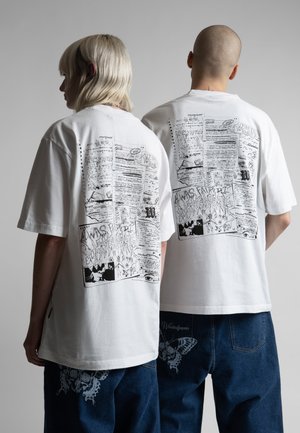 Δύο άνθρωποι στέκονται πλάτη με πλάτη φορώντας λευκά, μεγάλο μέγεθος t-shirt με μαύρα γραφικά κείμενα και σχέδια στην πλάτη, συνδυασμένα με μπλε τζιν.