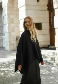 Svart poncho i mjukt tyg, med vid skärning och hög halsringning. Tillsammans med vita svarta byxor, mot en stenig bakgrund.