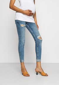 Jeans skinny azules desgastados con desgarros en las rodillas, puños doblados, combinados con mule de tacón bajo color beige claro. Top blanco ajustado sobre una barriga de embarazada.
