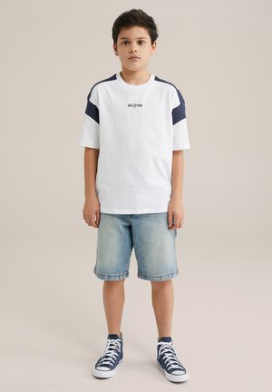 Witte t-shirt met korte mouwen en marineblauwe accenten, met de tekst "SALTYDOG", gecombineerd met lichtblauwe denim shorts en marineblauwe high-top sneakers.