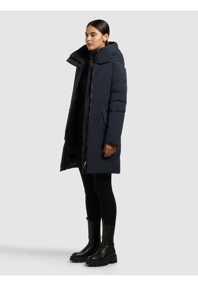 Navy plava puhasta jakna s visokim ovratnikom, s prednjim patentnim zatvaračem, bočnim džepovima i dugim krojem, uparena s crnim čizmama do koljena.