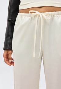 Κοντινό ενός ατόμου που φοράει ανοιχτό μπεζ παντελόνι με κορδόνι και μαύρο μακρυμάνικο τοπ με κοντό τελείωμα.