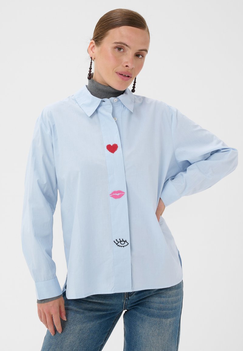 Femme portant une chemise bleu clair avec un cœur rouge, des lèvres roses et des motifs d'yeux noirs sur le devant, associée à un jean bleu et des boucles d'oreilles foncées.