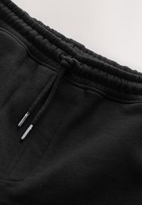 Gros plan de la ceinture d’un pantalon de jogging noir avec bande élastique et cordon noué avec embouts métalliques.