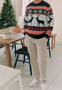 Strickpullover in Schwarz, Rot und Blau mit Rentier- und Schneeflockenmuster; kombiniert mit cremefarbenen Hosen und weißen Turnschuhen.