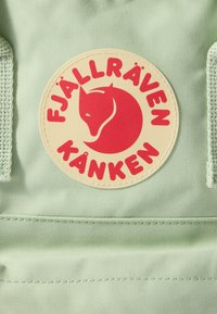 Fjällräven Kånken mugursoma ir izgatavota no gaiši zaļas auduma ar izteiksmīgu apaļu logotipu, kurā attēlots sarkans lapsas dizains uz bēša fona.