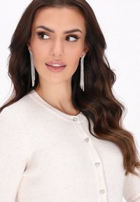 Rippenshirt in Creme mit herzförmigen Strassknöpfen; das Modell trägt lange, funkelnde silberne Ohrringe. Weicher Stoff, schlichtes Design.