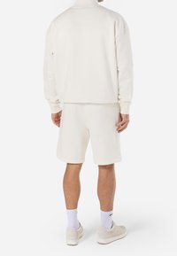 Ensemble sweat-shirt et short en polaire blanche. Coupe ample, col montant, poignets côtelés et texture douce. Porté avec des baskets blanches et des chaussettes.