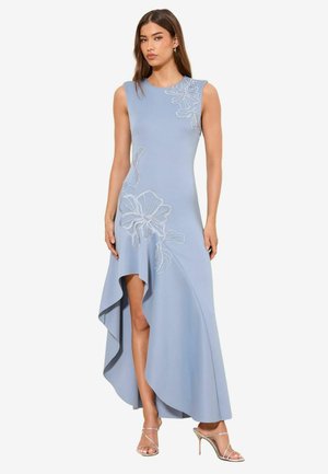 Kobieta w bezrękawnikowej, jasnoniebieskiej sukience o asymetrycznym kroju z kwiatowym haftem, w srebrnych sandałkach na obcasie, stoi na białym tle.