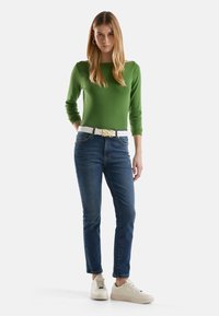 Groene aansluitende trui, blauwe jeans en een witte riem met gouden accenten. Model draagt crèmekleurige sneakers en staat tegen een neutrale achtergrond.