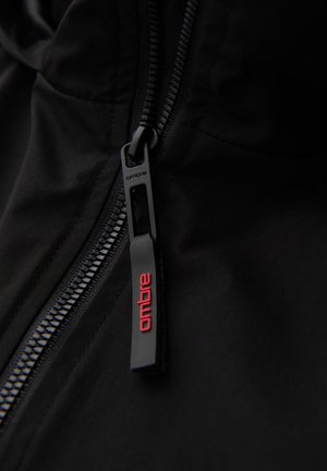 Sort zip-jakke med detaljer som en glat lynlås med et mærket trækstykke i mat sort og røde accenter. Tekstureret stof er synligt.