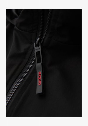 Détail d'une veste zippée noire présentant une fermeture éclair lisse avec un tirette de marque en noir mat et des accents rouges. Tissu texturé visible.