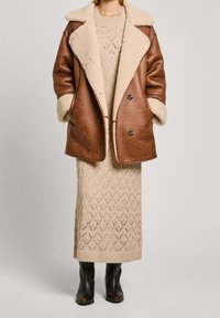 Kvinna iklädd en brun fårskinnsjacka över en beige lång klänning med öppen väv och svarta läderstövlar, stående mot en neutral bakgrund.