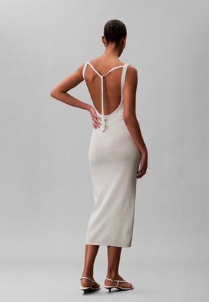 Mujer con vestido midi blanco, sin espalda y sin mangas, con tirantes finos atados detrás del cuello, de pie con una mano en la cadera y calzando sandalias de tacón bajo.