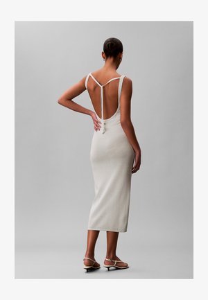 Frau in einem weißen, rückenfreien Midi-Kleid ohne Ärmel mit dünnen Trägern, die hinter dem Nacken gebunden sind, steht mit einer Hand in der Hüfte und trägt Sandalen mit niedrigem Absatz.