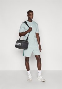 Αθλητική πράσινη ανοιχτόχρωμη μπλούζα και σορτς, που κρατούν μια μαύρη τσάντα Adidas με λευκό λογότυπο. Τα παπούτσια είναι λευκά με γκρι και εκρού λεπτομέρειες.