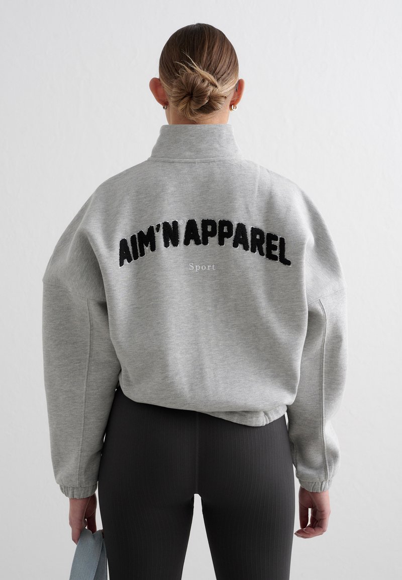 Grå genser med høy krage og oversized passform. Har svart brodert tekst med påskriften "AIM'N'APPAREL Sport" på baksiden.