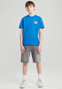 Sininen puuvillapaita lyhyillä hihoilla ja rintatasku, yhdistettynä harmaisiin farkkushortseihin ja valkoisiin tennareihin. Yksinkertainen tausta.