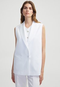 Witte mouwloze blazer met een notch-lapel, twee voorzakken en een enkele knoopsluiting. Gemaakt van een gladde, lichte stof.