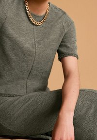 Zielony, teksturowany strój z krótkimi rękawami, z okrągłym dekoltem i podwiniętymi rękawami. Złoty naszyjnik z łańcuszka stanowi kontrastujący detal.