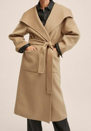 Mujer con un abrigo cruzado beige con gran cuello, cintura ceñida con cinturón, bolsillos aplicados, sobre una camisa negra abotonada y pantalones negros.
