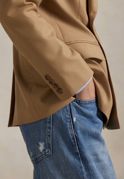 Béžový blazer so štyrmi hnedými gombíkmi na rukáve, skombinovaný s opotrebovanými modrými džínami. Materiál vyzerá hladko a má klasický dizajn.