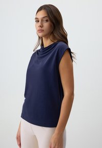 Donna con lunghi capelli castani, vestita con un top senza maniche di colore blu navy con scollo drappeggiato e pantaloni beige chiaro, in piedi su uno sfondo chiaro e semplice.